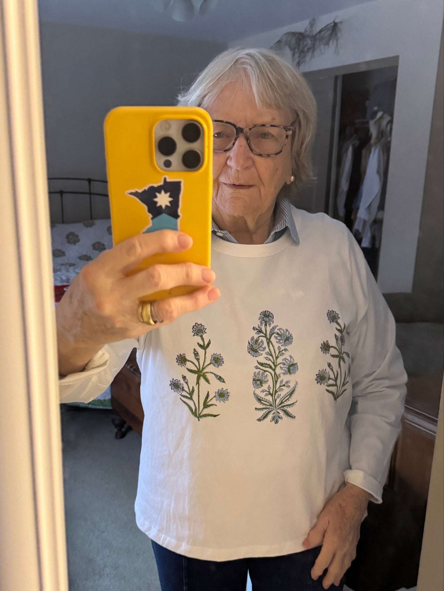 Nonie taking a mirror selfie with a yellow phone, wearing a white shirt with floral patterns.