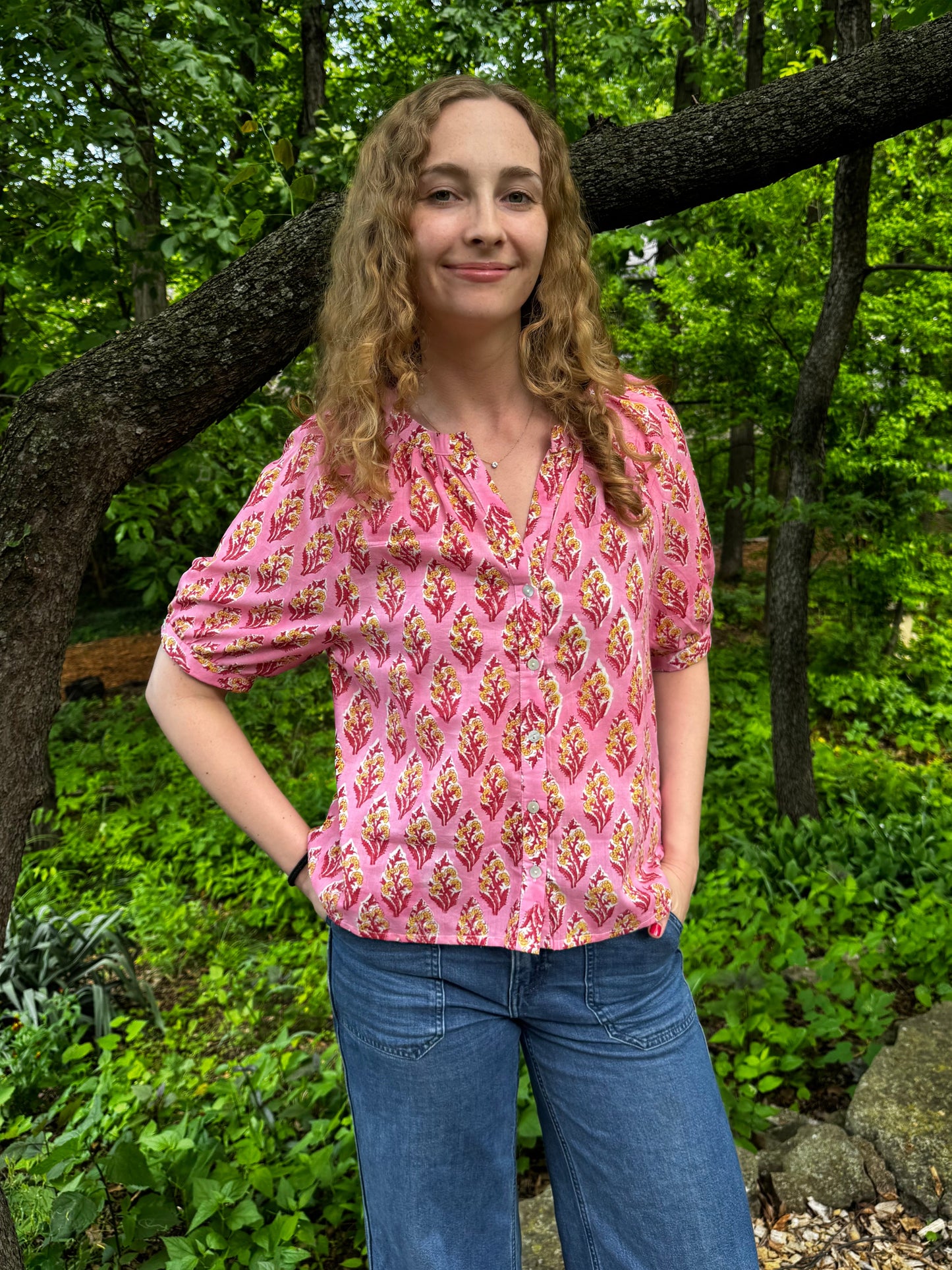 Woman in a pink patterned blouse and blue jeans standing in a forest