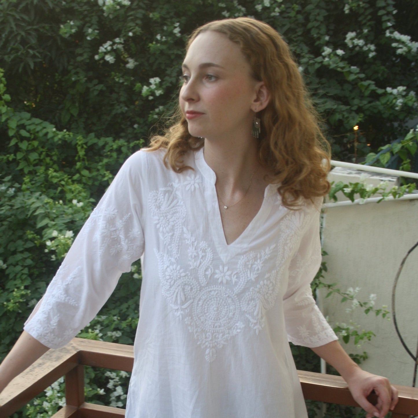 A woman standing on a balcony wearing a white cotton tunic with hand-stitched Chikankari embroidery, three-quarter length sleeves, a v-neckline, a band collar, and thigh-length hem with side slits.