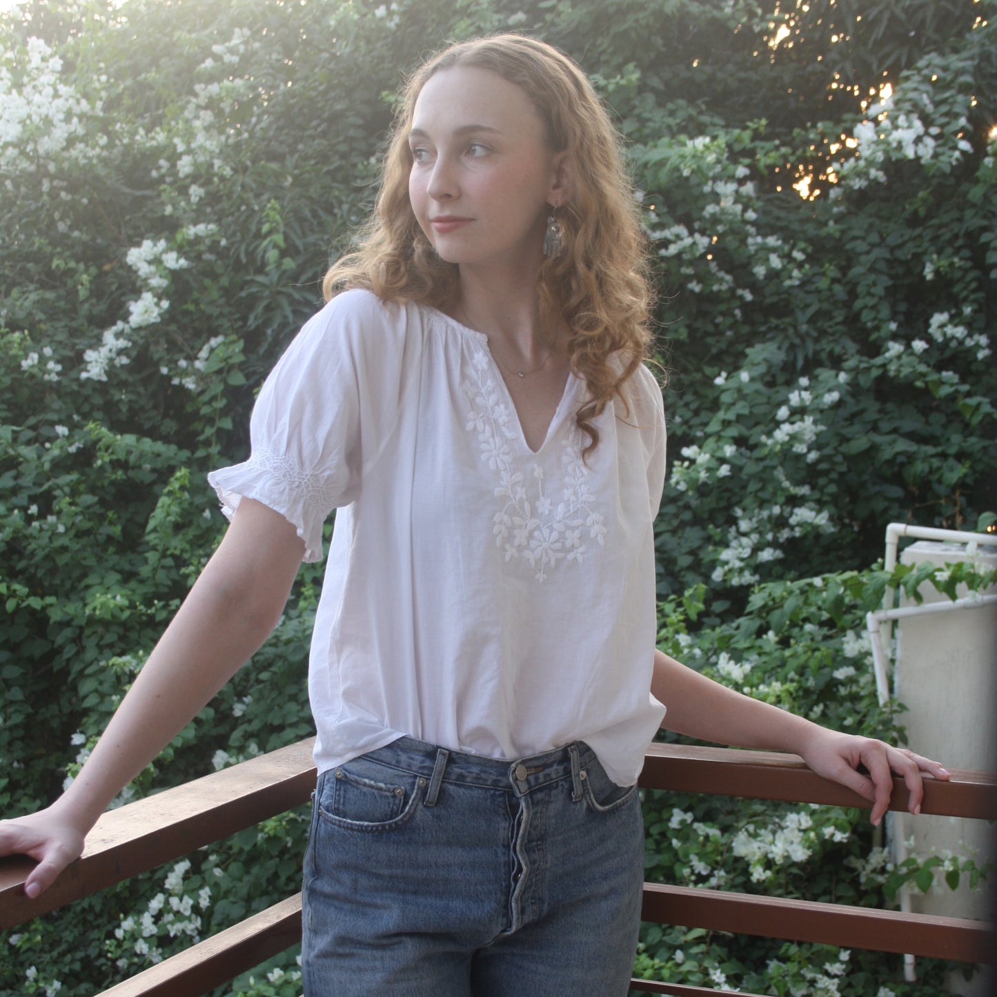 Person wearing a white blouse and blue jeans standing outdoors with greenery in the background