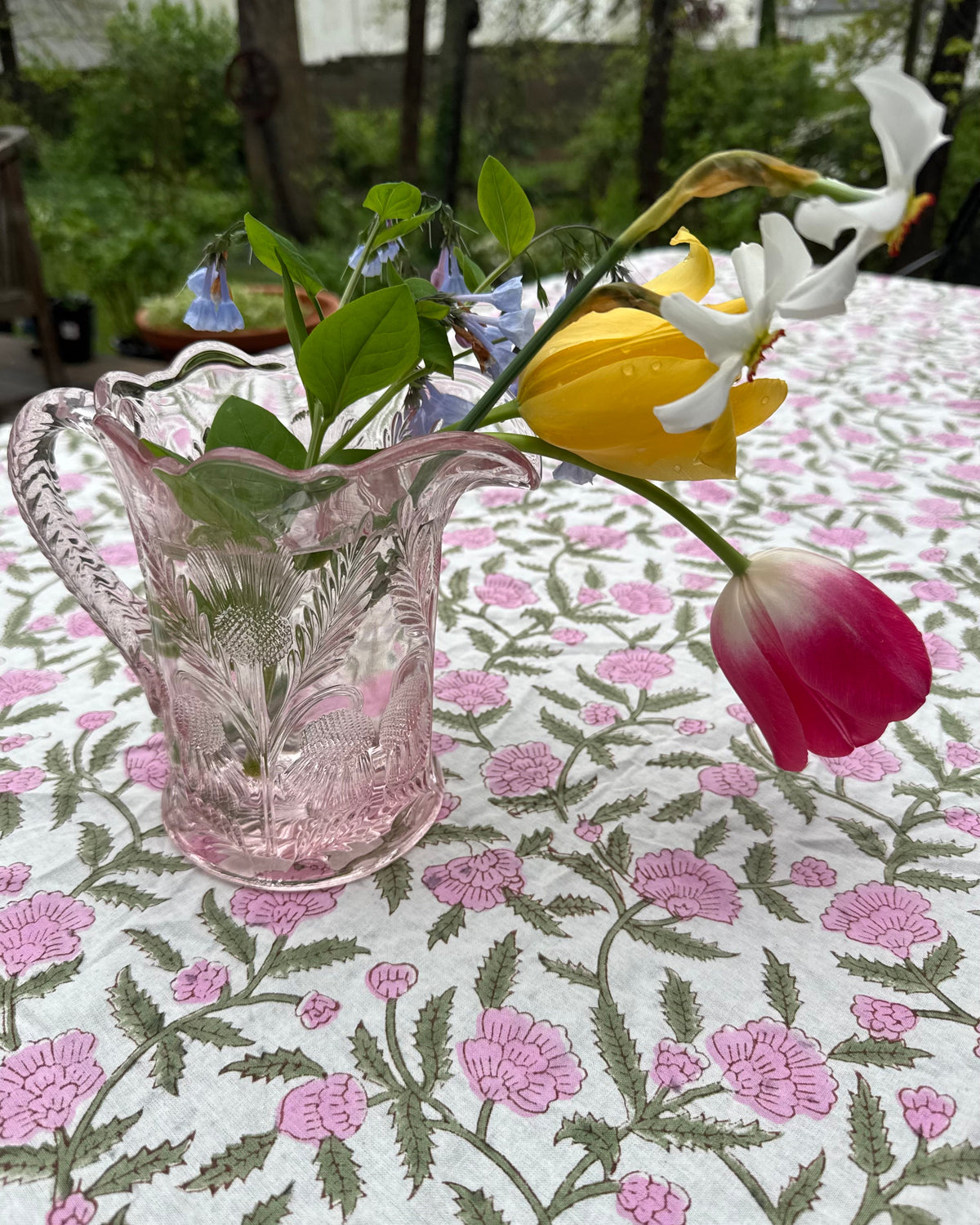 A pink glass pitcher filled with tulips and small blue flowers, placed on a white tablecloth with a pink floral pattern. The background shows an outdoor garden setting.