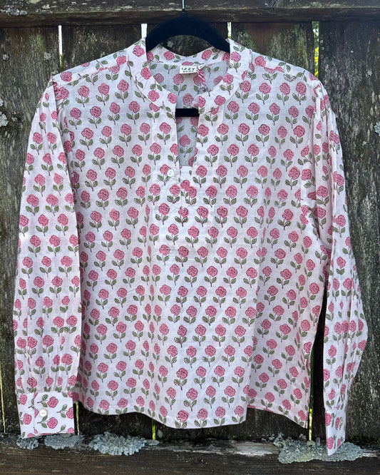 A long-sleeve button-up blouse with a pink and green floral block print on a white base, hanging on a black hanger against a rustic wooden fence.