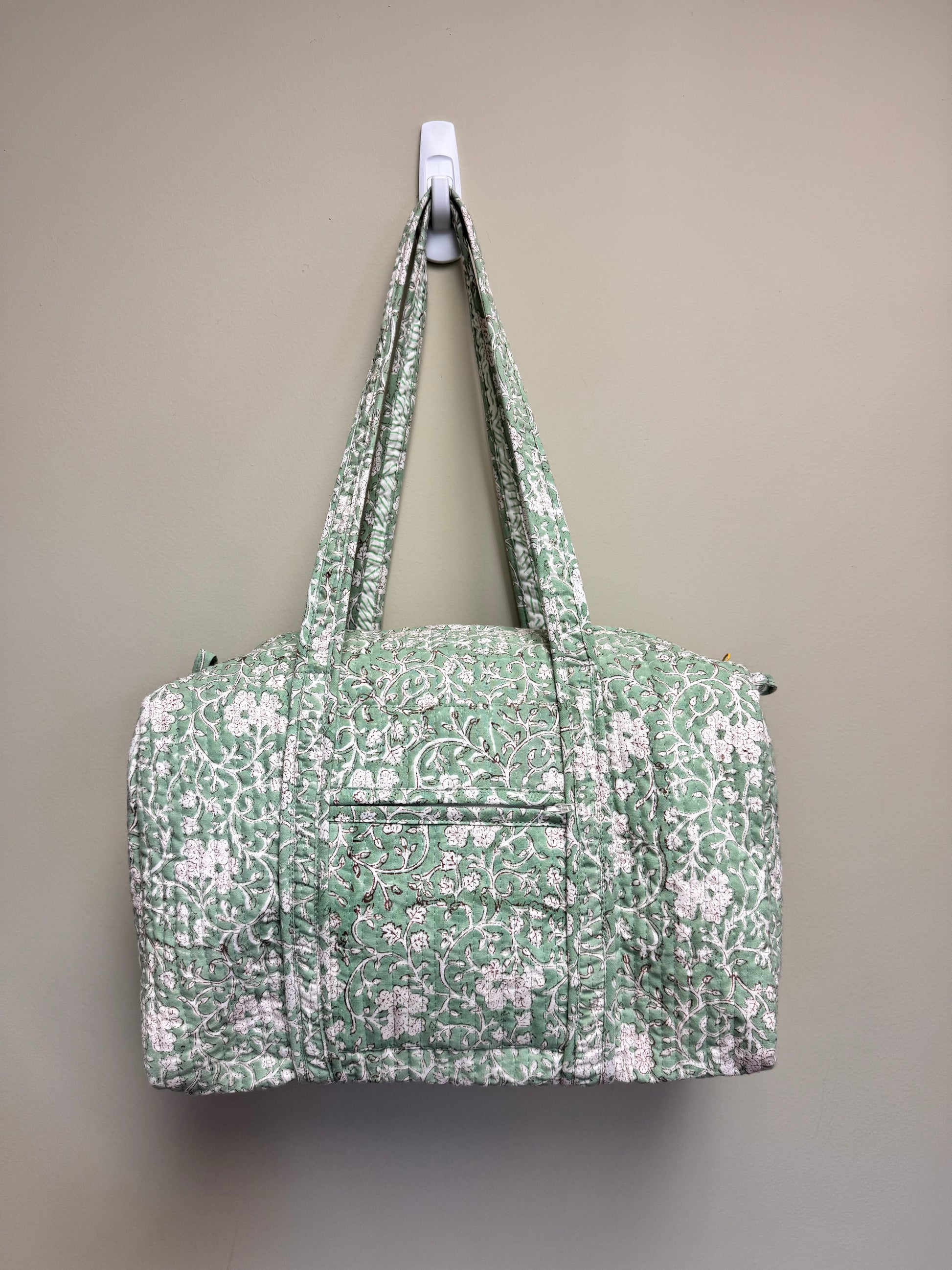 A quilted duffel bag with a green and white floral pattern, hanging from a white wall hook against a solid beige wall.