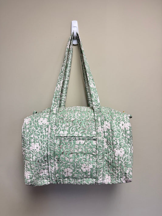 A quilted duffel bag with a green and white floral pattern, hanging from a white wall hook against a solid beige wall.