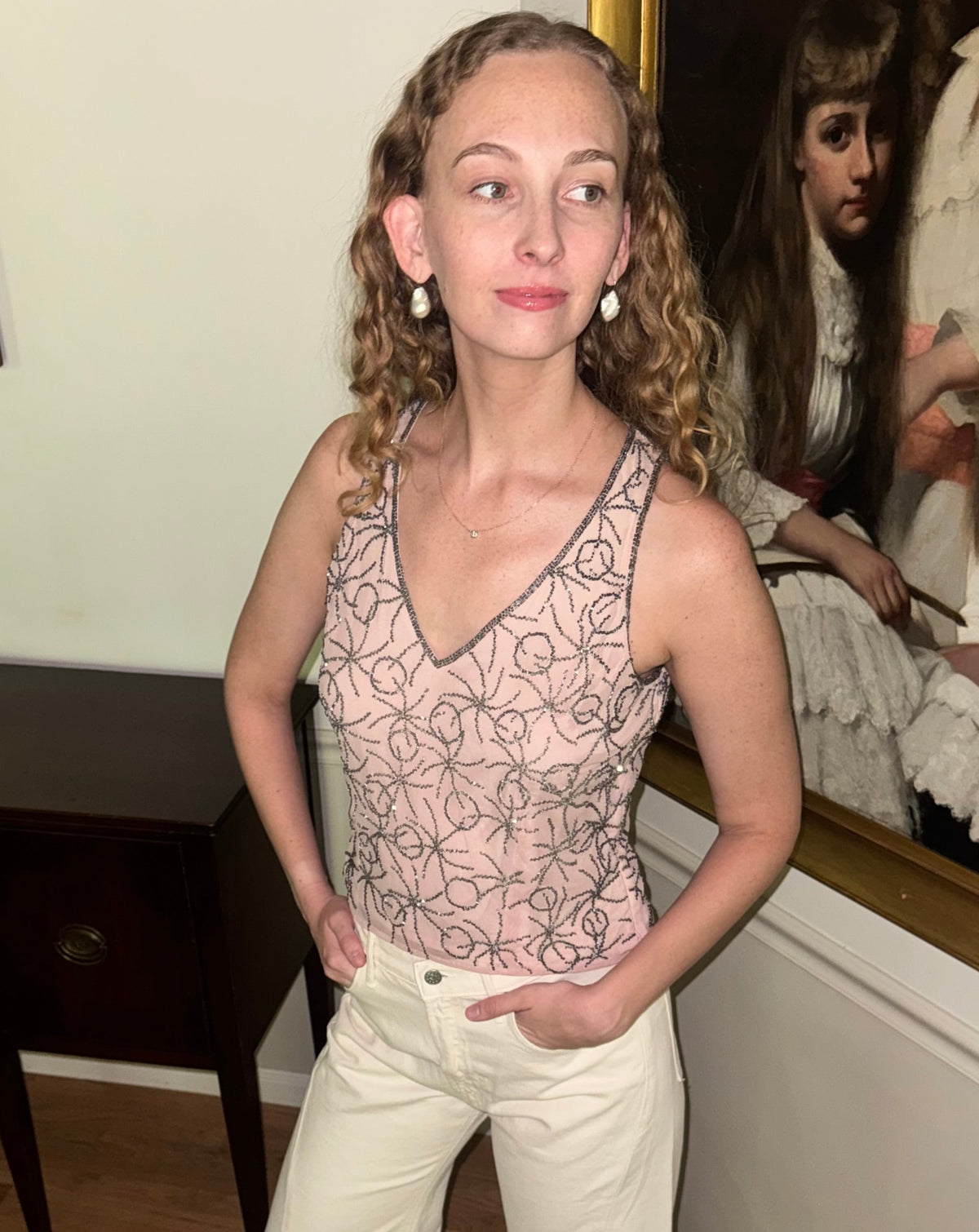 A woman stands indoors wearing a sleeveless lace bodysuit with a floral pattern in pale pink and cream, paired with cream wide-leg pants. She is posed in front of a wall with a large framed portrait and a dark wooden side table.