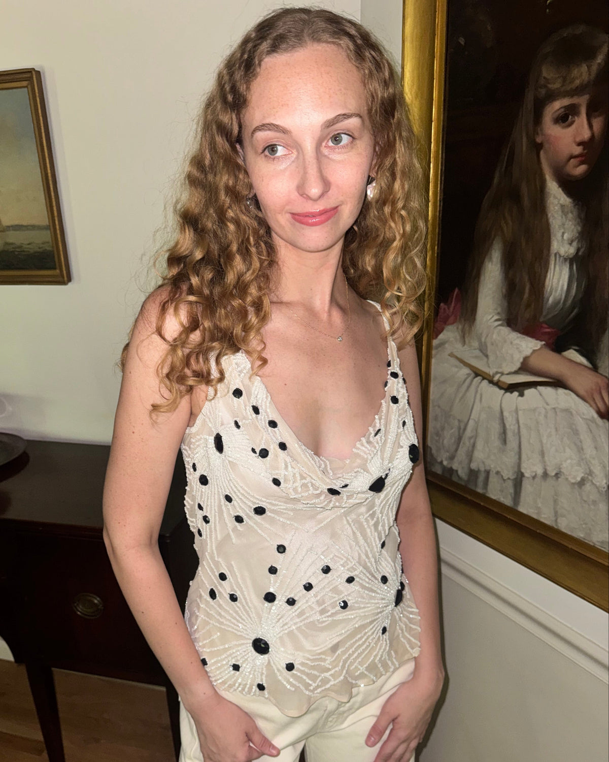 A woman with long curly light brown hair stands indoors wearing a cream lace sleeveless top with black polka dots and matching cream pants. She is photographed in a room with wood flooring, framed wall art, and a dark wooden table.