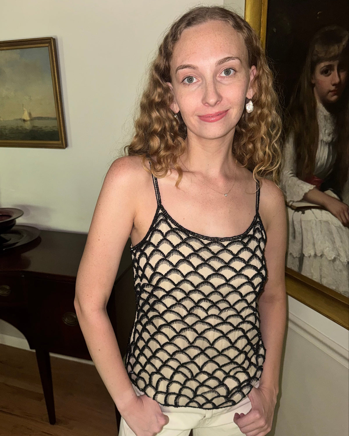 A woman with curly blonde hair wearing a black fish-scale patterned crochet tank top and cream pants, standing in an indoor room with framed art and a mirror in the background.