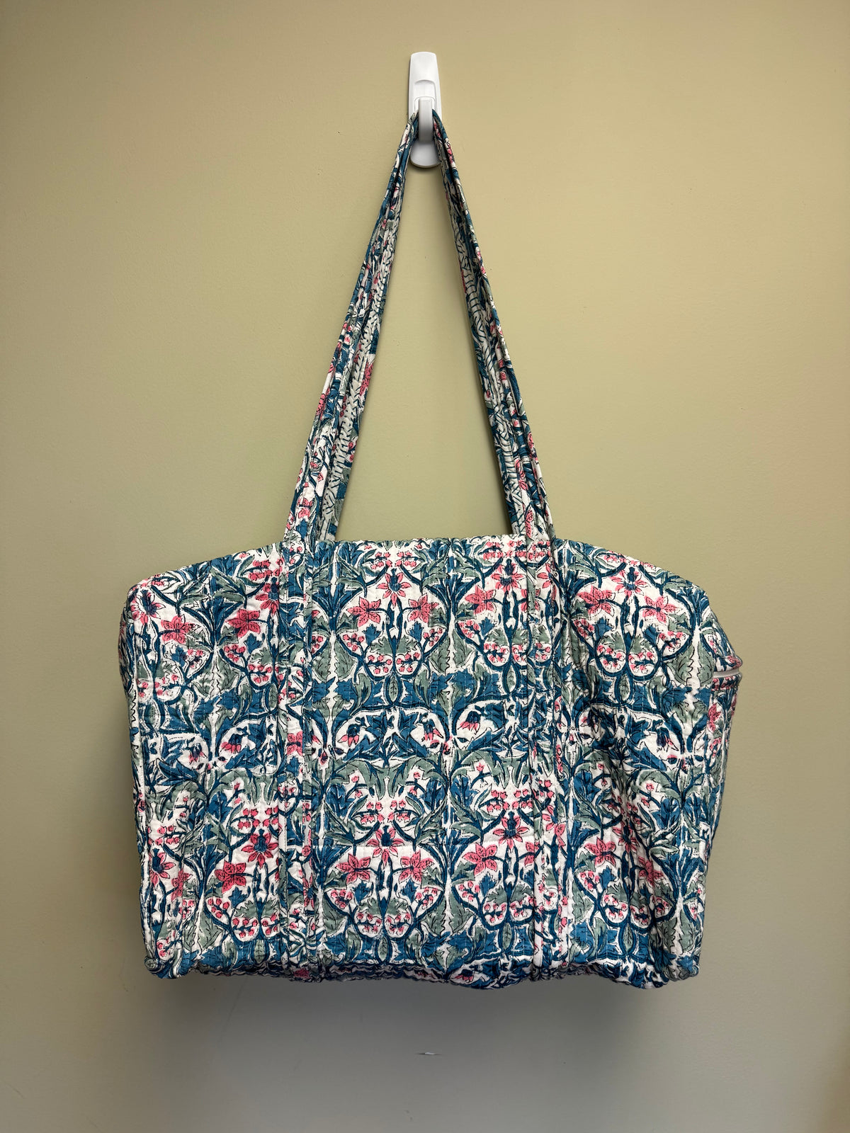 A large tote bag with a blue, pink, and white floral damask pattern, hanging from a white plastic hook against a beige wall.