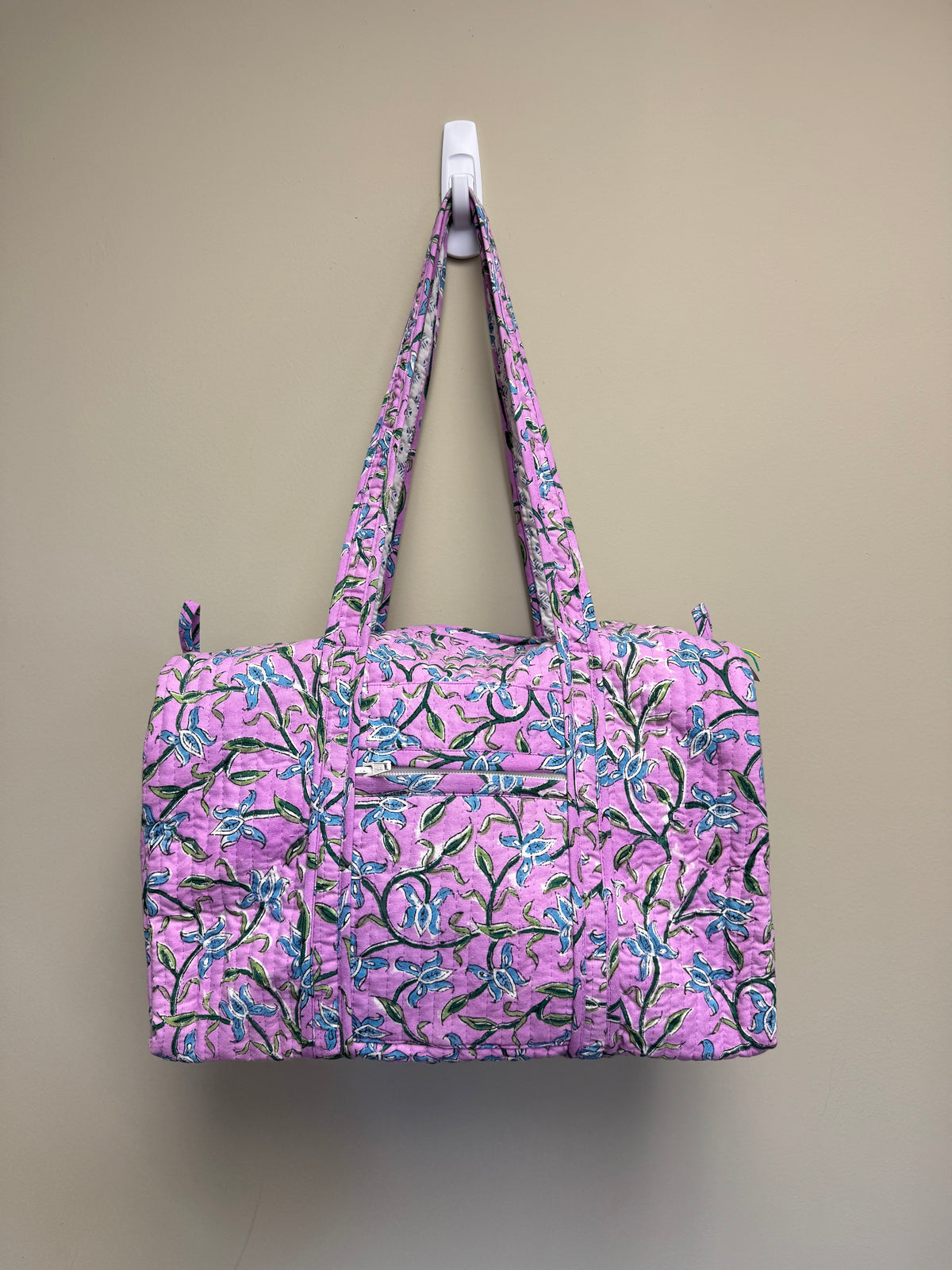 A quilted duffel bag with a pink and purple floral pattern, hanging from a white wall hook against a beige wall.