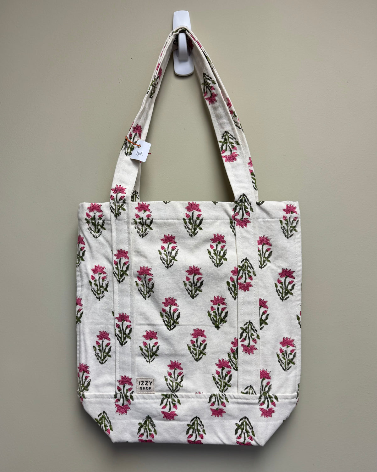 A white canvas tote bag with a repeating pink and green floral block print, hanging from a white wall hook against a neutral beige wall.