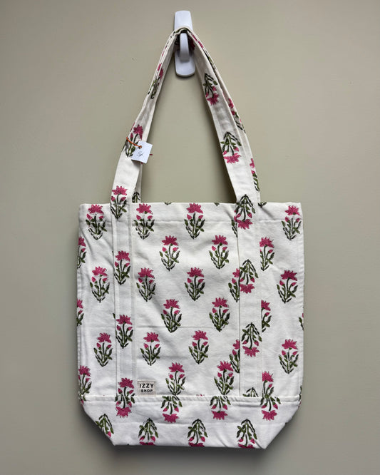A white canvas tote bag with a repeating pink and green floral block print, hanging from a white wall hook against a neutral beige wall.