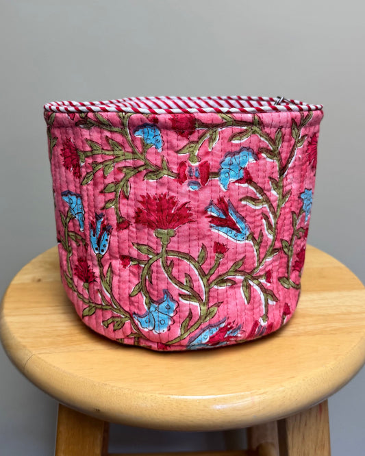 A pink quilted fabric storage basket with a floral pattern sits on a round wooden stool against a plain gray wall.