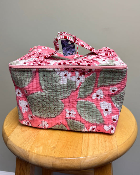 A quilted fabric storage bag with a pink and green floral pattern sits on a round wooden stool against a plain gray wall.