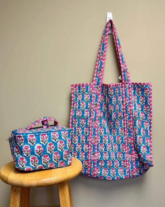 A large tote bag and a matching small lunch bag with a pink and blue floral block print, displayed against a beige wall with one hanging on a hook and one resting on a wooden stool.