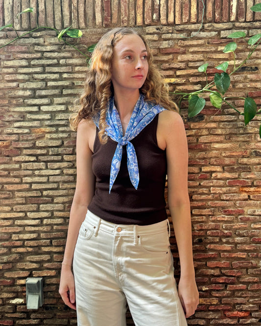 A woman with long wavy light brown hair stands against a rustic brick wall with green vines, wearing a dark brown sleeveless top, cream-colored pants, and a blue patterned neck scarf.