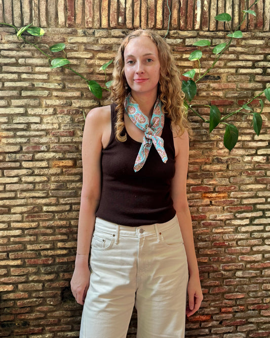 A woman with curly blonde hair stands against a rustic brick wall with green vines, wearing a dark brown sleeveless top, cream-colored pants, and a colorful paisley neck scarf.
