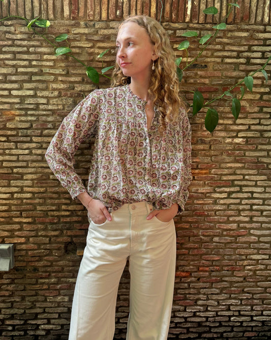 A woman with curly blonde hair stands against a rustic brick wall wearing a long-sleeve button-up blouse with a dense red, green, and beige floral pattern, paired with cream-colored wide-leg trousers.
