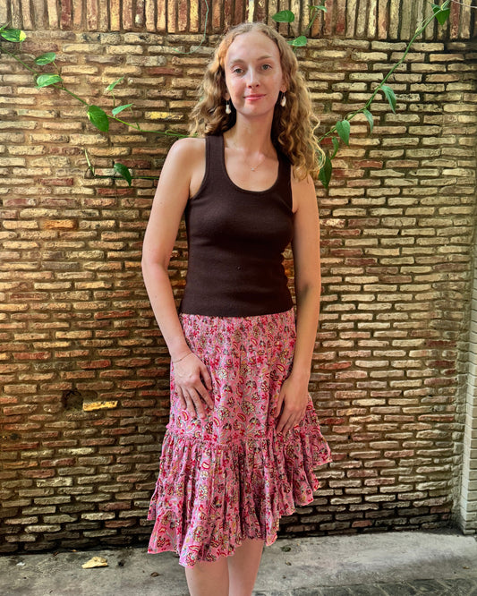 A woman stands in front of a rustic brick wall with green vines, wearing a dark brown ribbed tank top and a pink floral tiered ruffle skirt.