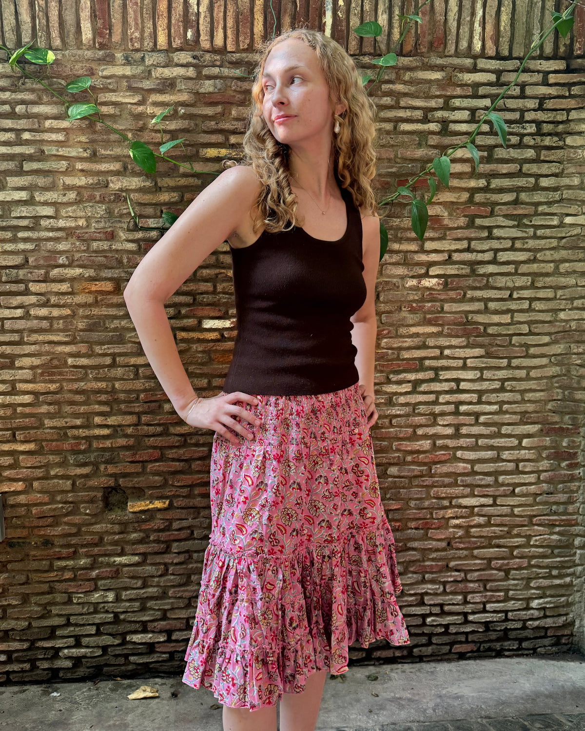 A woman stands in front of a brick wall wearing a dark brown ribbed tank top and a pink floral tiered midi skirt.