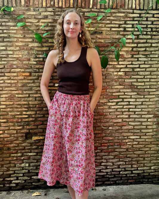 A woman stands in front of a rustic brick wall with green vines, wearing a dark brown ribbed tank top and a pink floral midi skirt with pockets.