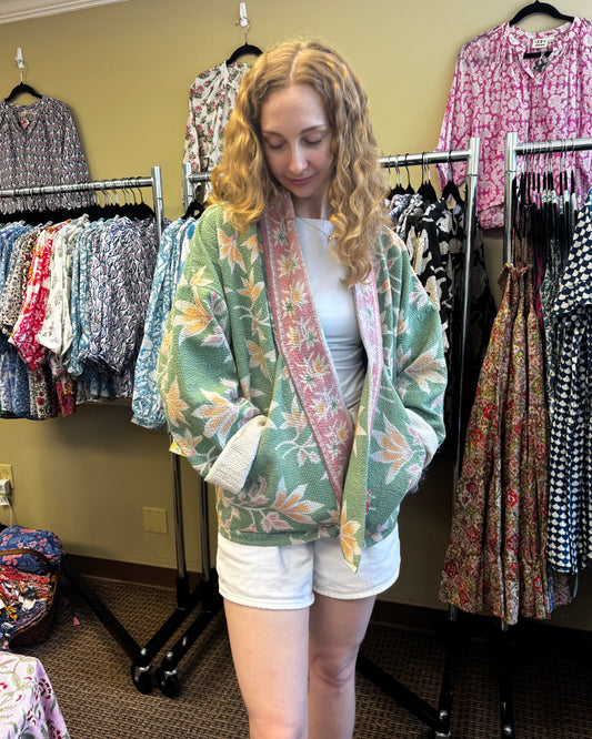 A woman with curly blonde hair stands in a clothing boutique wearing a sage green floral kimono-style cardigan with a pink lining over a white tank top and white shorts. Clothing racks filled with various patterned garments are visible behind her against a beige wall.