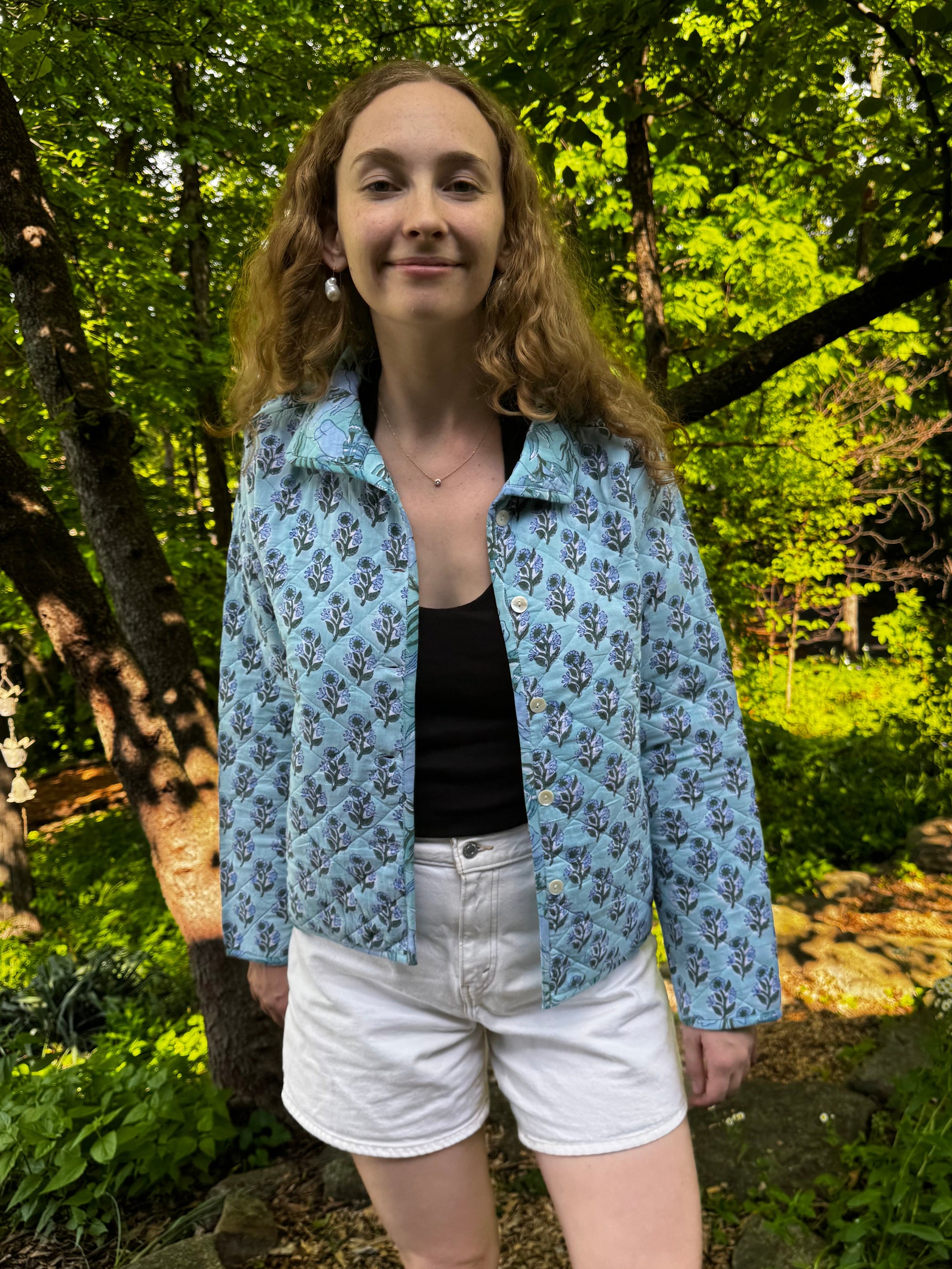 Woman standing outdoors in a forested area wearing a blue floral jacket.