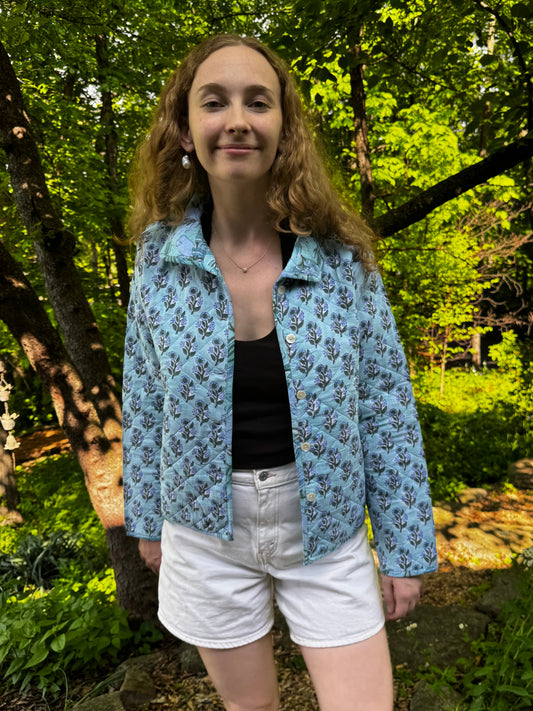 Woman standing outdoors in a forested area wearing a blue floral jacket.