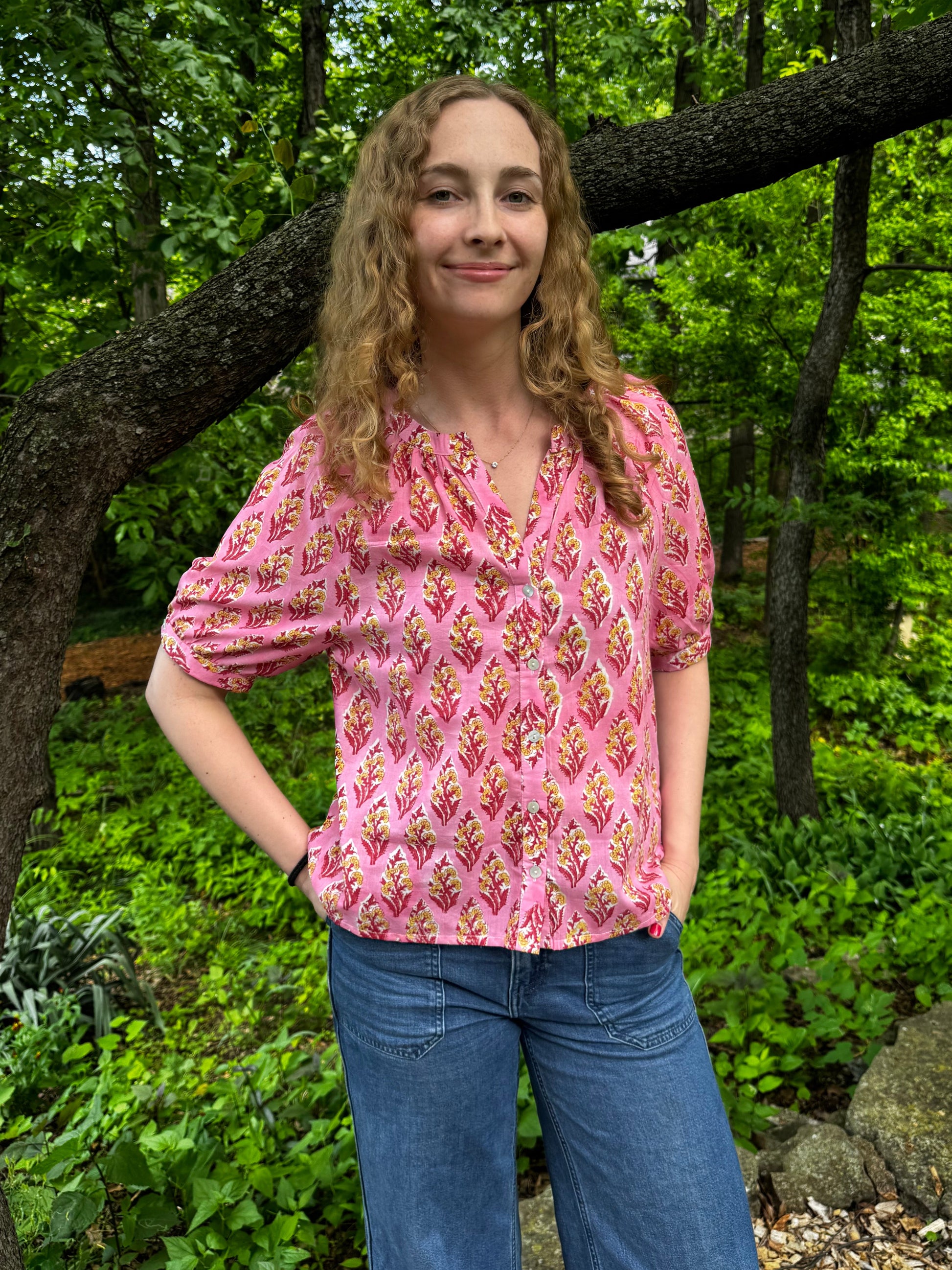 Woman in a pink patterned blouse and blue jeans standing in a forest
