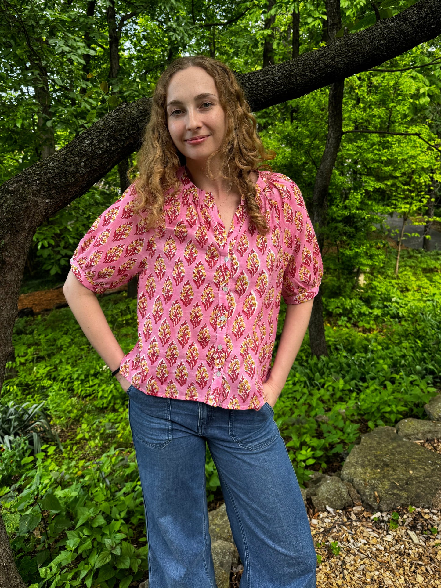 Woman in a pink patterned blouse and blue jeans standing in a forested area.