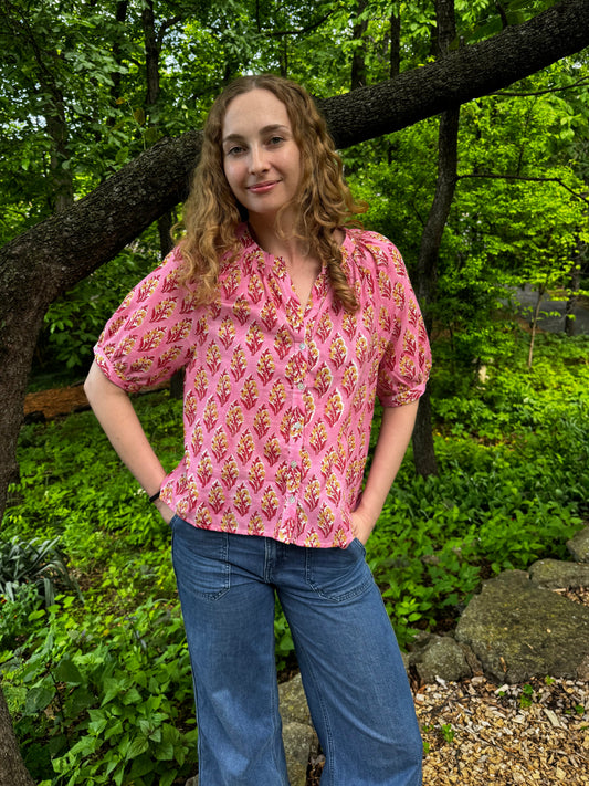 Woman in a pink patterned blouse and blue jeans standing in a forested area.