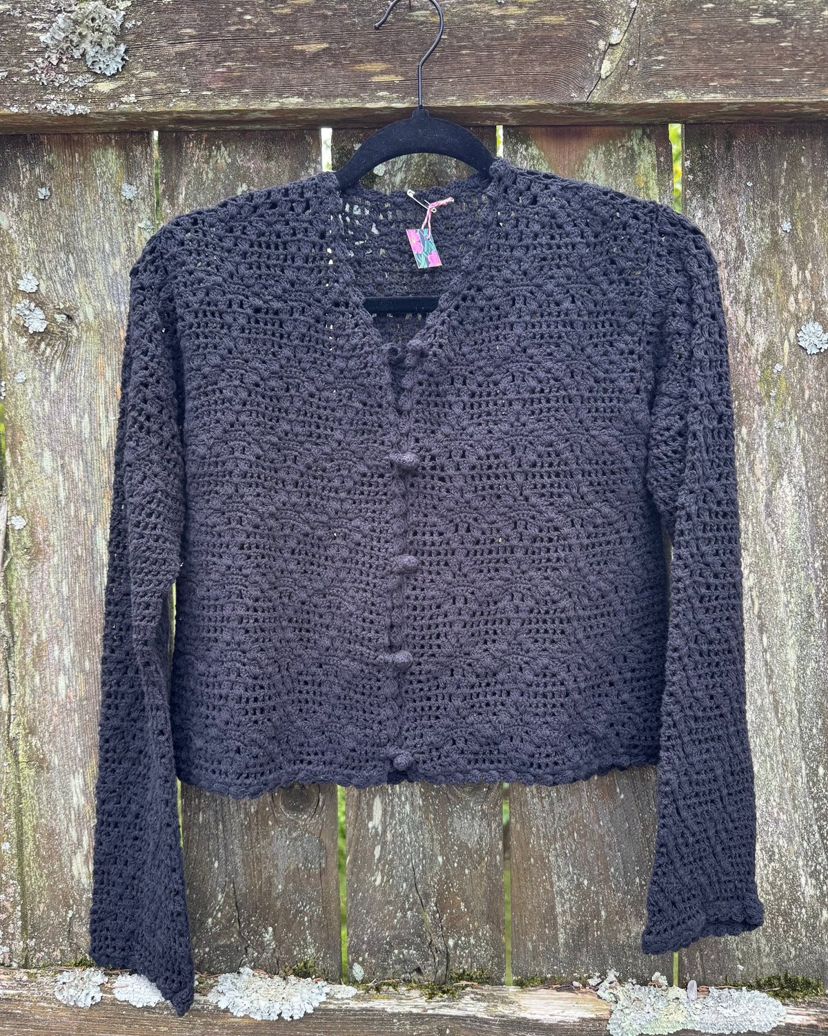 A black open-knit cardigan sweater with a button-up front, hanging on a black hanger against a weathered wooden fence background.