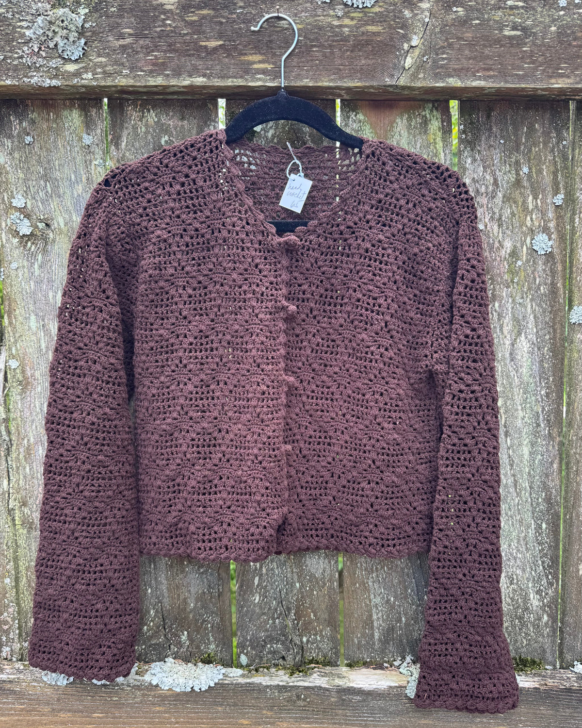 A brown crochet cardigan sweater with long sleeves and a V-neck, hanging on a black hanger against a weathered wooden fence.