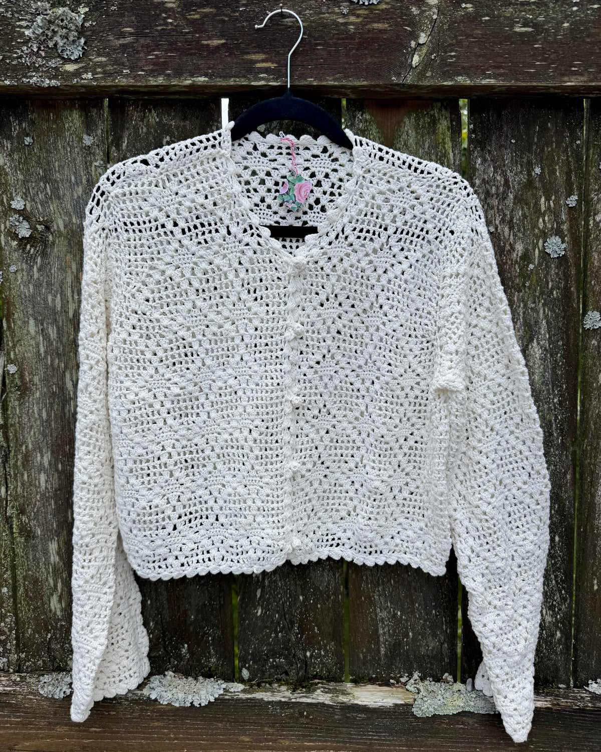 A white crochet cardigan sweater with a scalloped edge and button closure, hanging on a black hanger against a rustic weathered wooden fence.