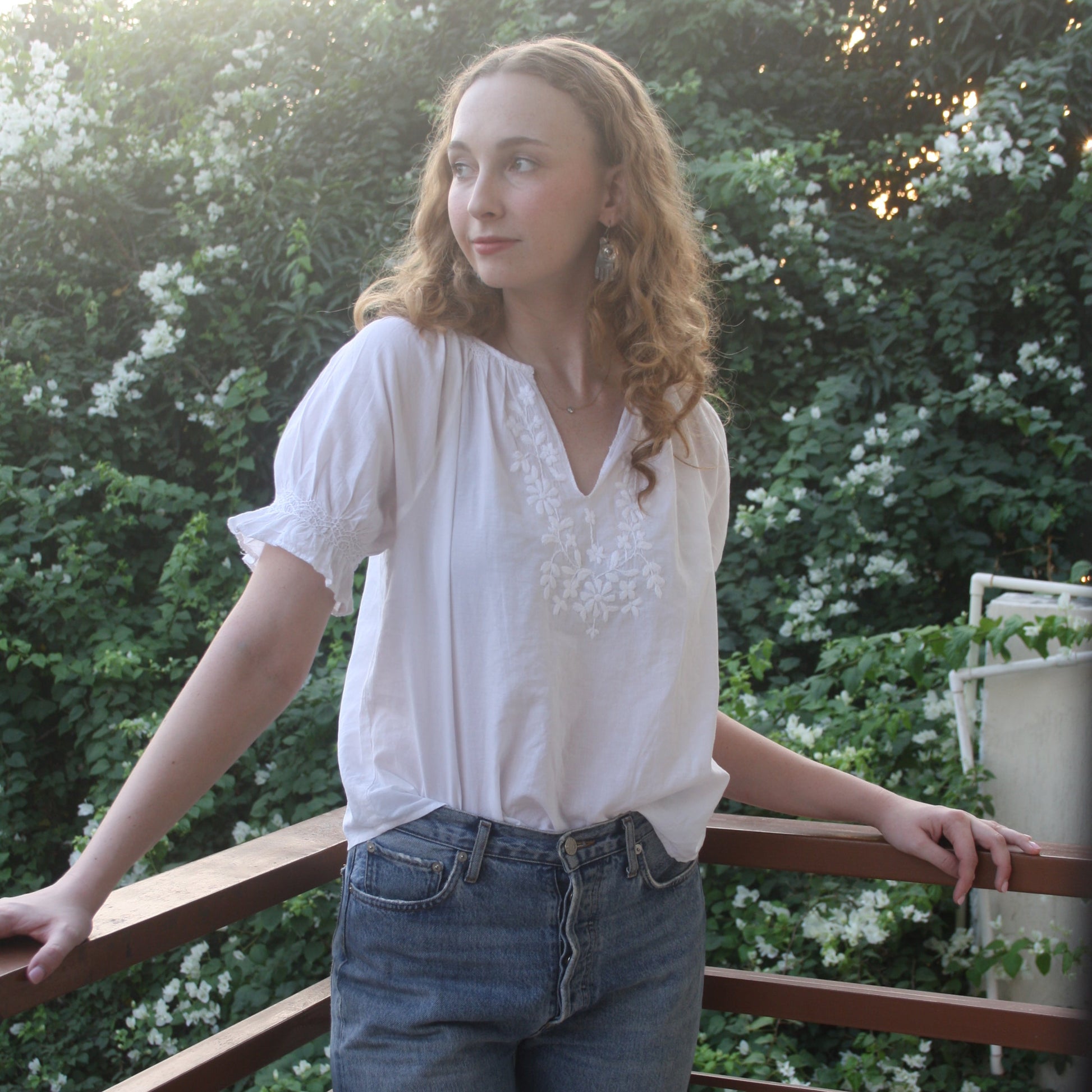 Person wearing a white blouse and blue jeans standing outdoors with greenery in the background